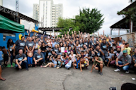 A large, diverse group of people, including adults and children, gathered outdoors, smiling and raising their hands in celebration. Urban buildings and greenery are visible in the background, creating a sense of community and solidarity.
